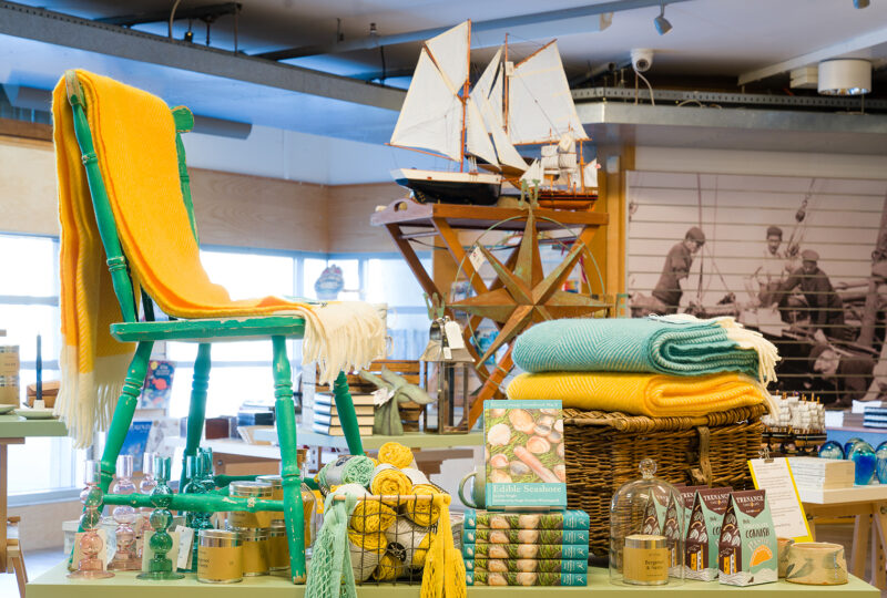 A photograph of a nautical shop display, including turquoise and yellow coloured throws, model boats and piles of books.