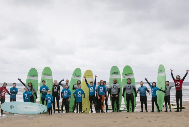 A photograph of a group of children and adults standing on the beach in front of their surfboards, which are standing upright in the sand.