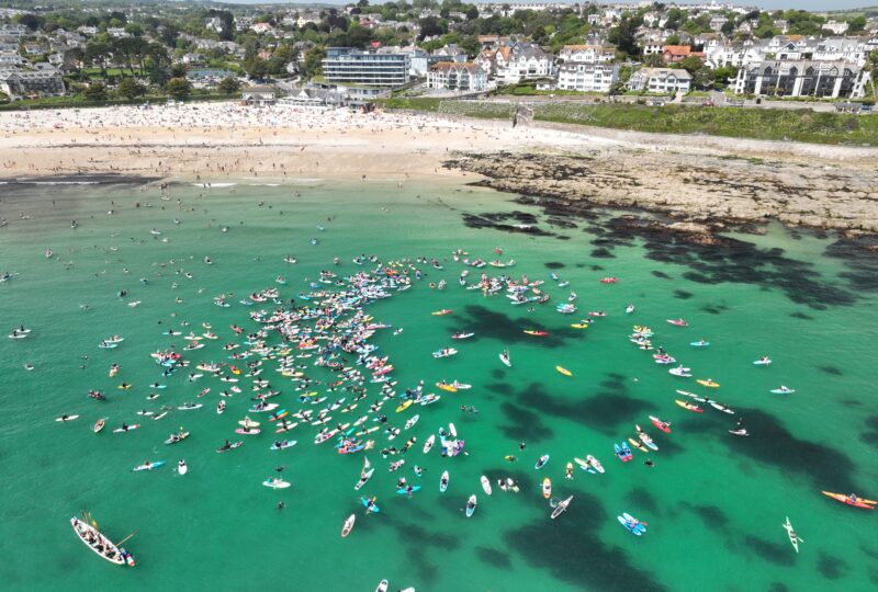 An aerial photograph taken above a beach showing hundreds of colourful surfboards and paddleboards gathered together in the sea.