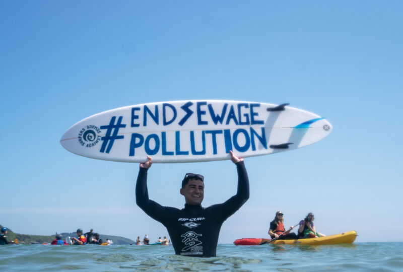 A photograph of a man standing in the sea wearing a black wetsuit, holding a surfboard horizontally above his head - the surfboard has the words '#End Sewage Pollution' painted on it.