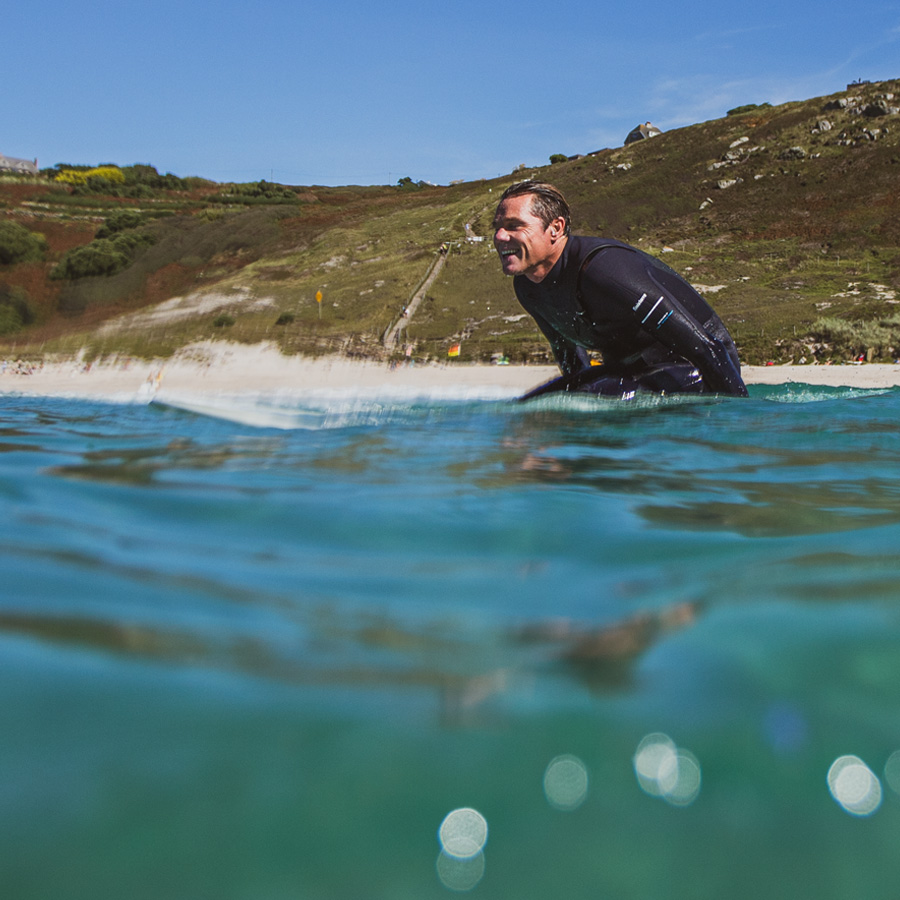 A photograph of a smiling surfer in a wetsuit sitting on their board, with a sunny sky and landscape behind them as they look out to sea.