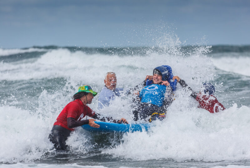 A photograph showing someone on an adapted surfboard being supported by three men standing in the water.