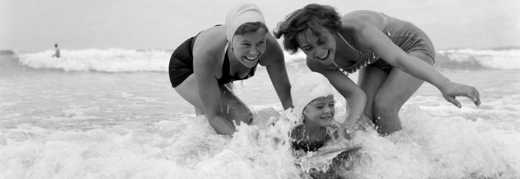 A black and white photograph of Two women in swimsuits play in the ocean waves, helping a person riding a surfboard, capturing a moment of fun at the beach.
