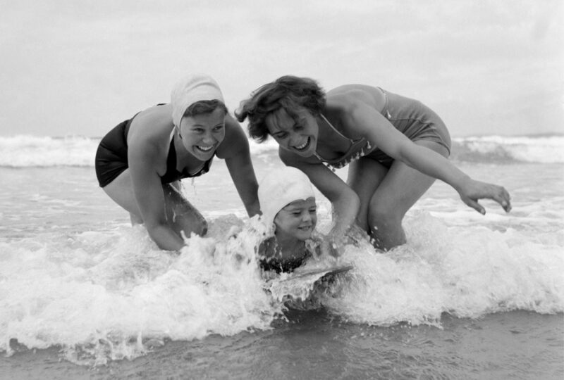 A black and white photograph of Two women in swimsuits play in the ocean waves, helping a person riding a surfboard, capturing a moment of fun at the beach.