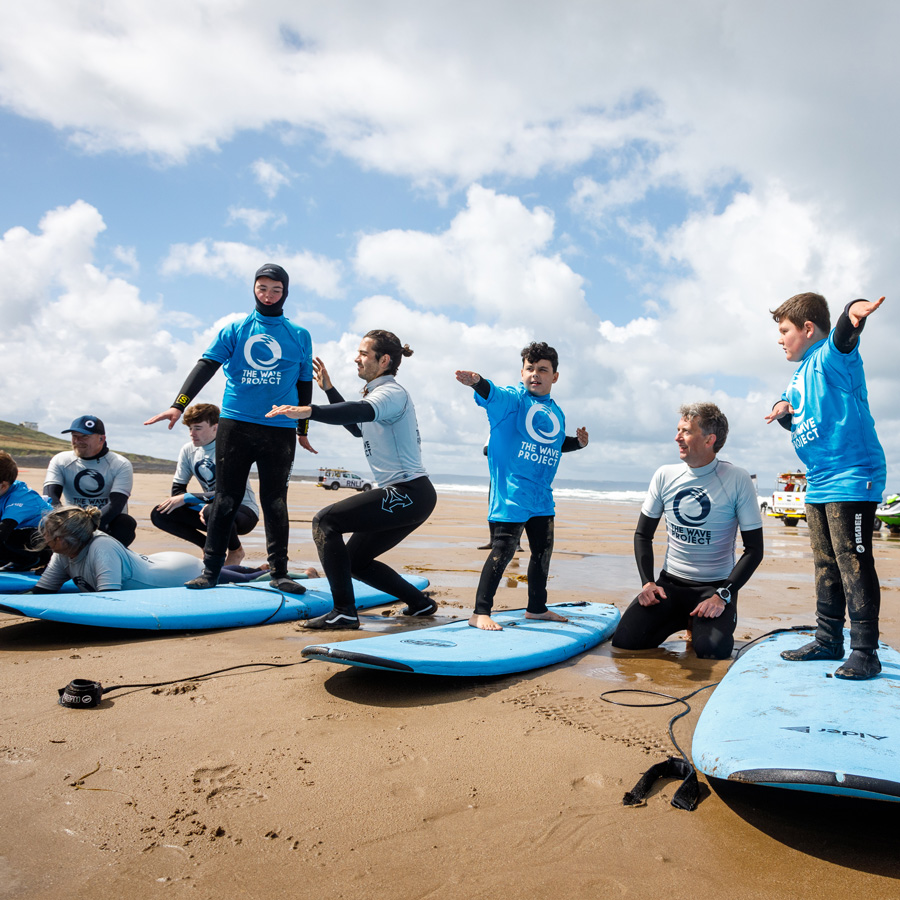 A photograph showing a group of people on a beach being taught how to stand on their surfboards, learning to surf.