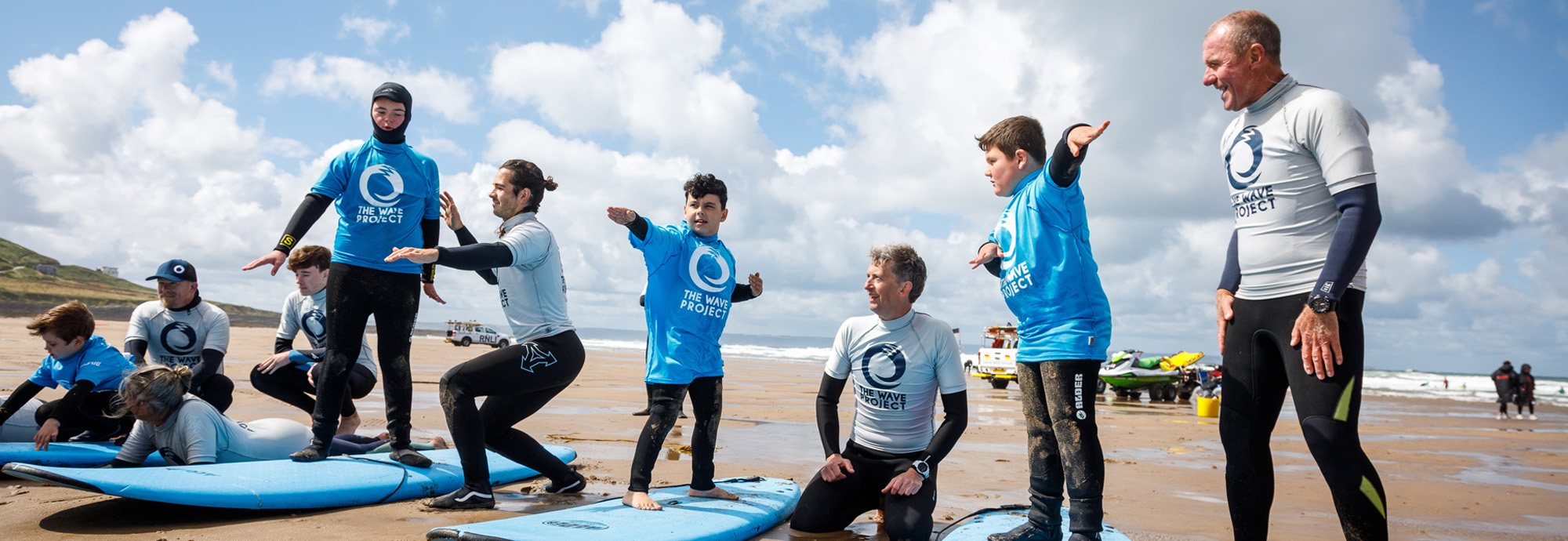 A photograph showing a group of people on a beach being taught how to stand on their surfboards, learning to surf.