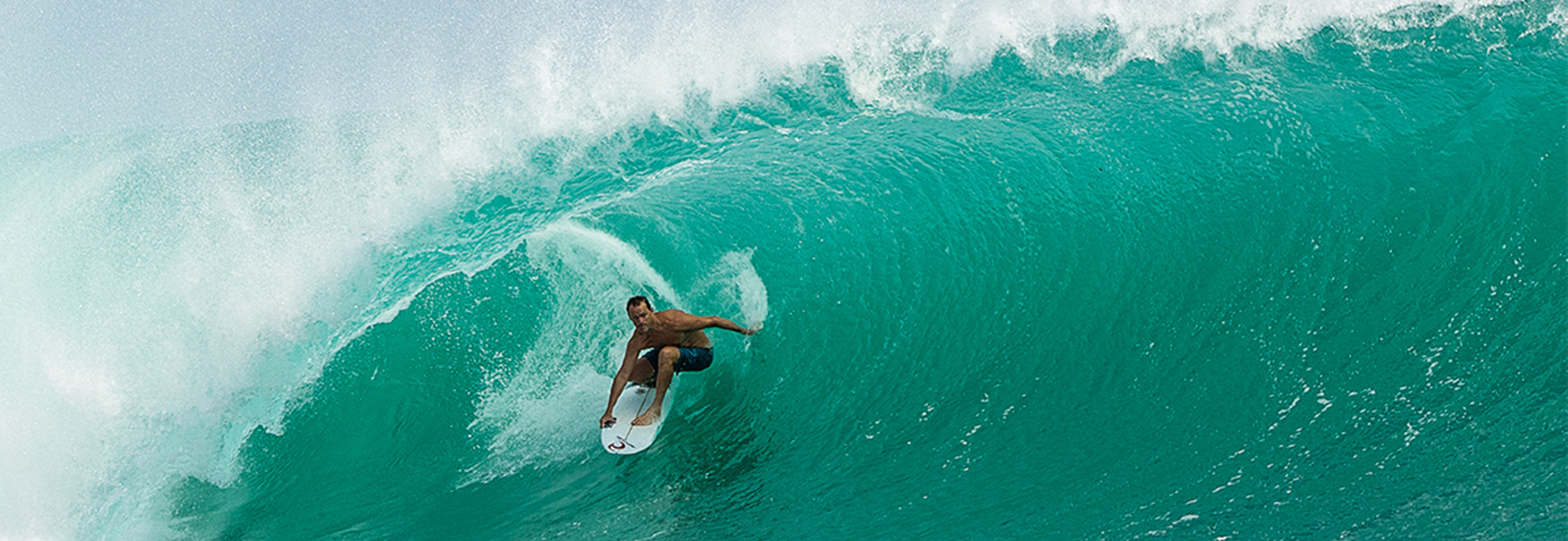 A photograph of a male surfer riding the barrel of a wave while a photographer uses a large camera from the water.