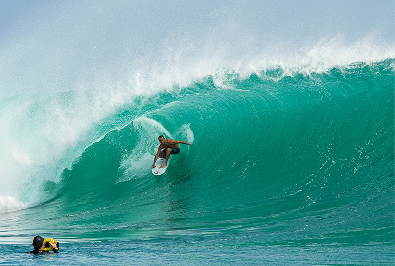 A photograph of a male surfer riding the barrel of a wave while a photographer uses a large camera from the water.