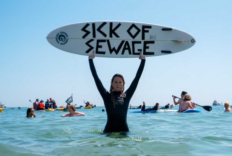 A photograph shows a person wearing a wetsuit, standing in the sea, holding a board above their head that reads 'Sick of Sewage'.