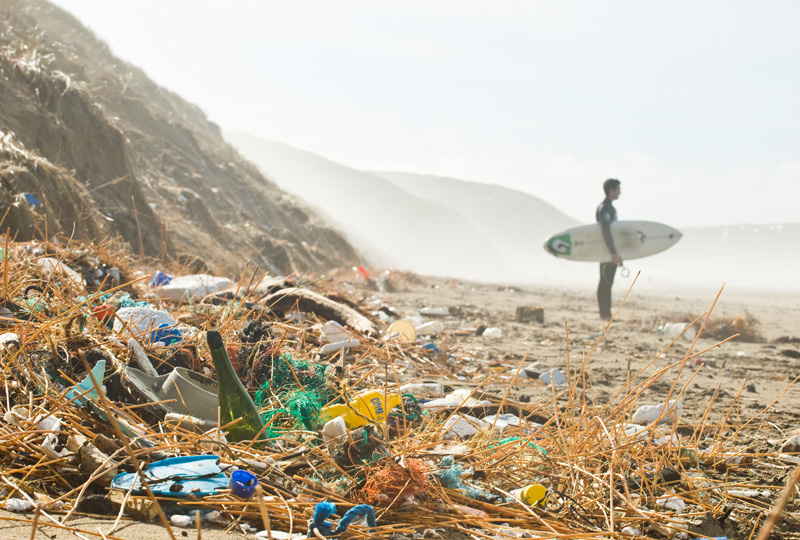 A photograph of a beach, showing piles of waster on the shoreline in the foreground, and a surfer holding their board in the background.