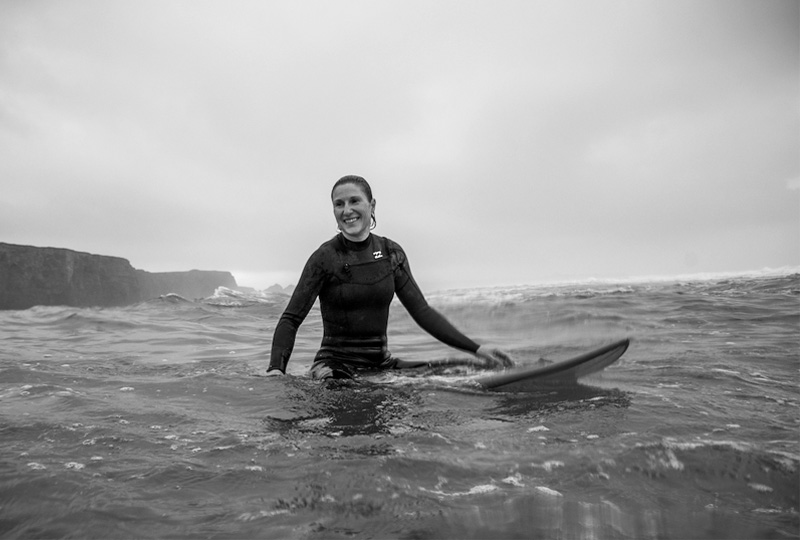 A black and white photograph showing a female surfer sitting on their board in the water, smiling.