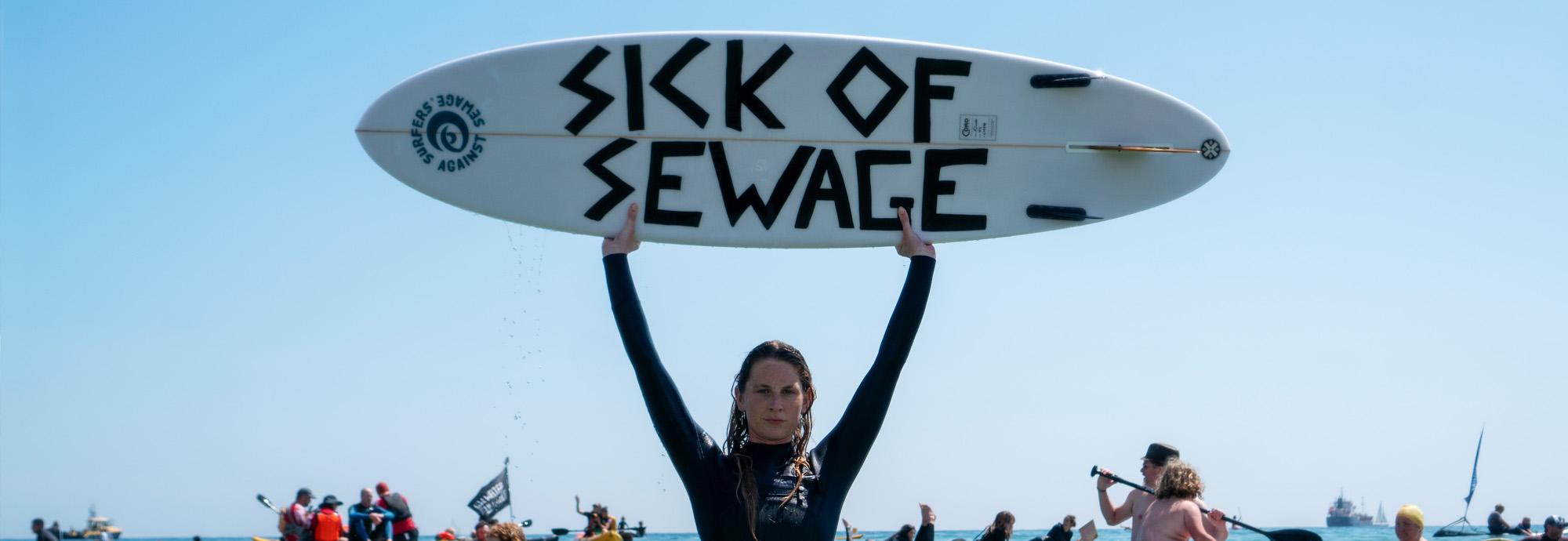 A photograph shows a person wearing a wetsuit, standing in the sea, holding a board above their head that reads 'Sick of Sewage'