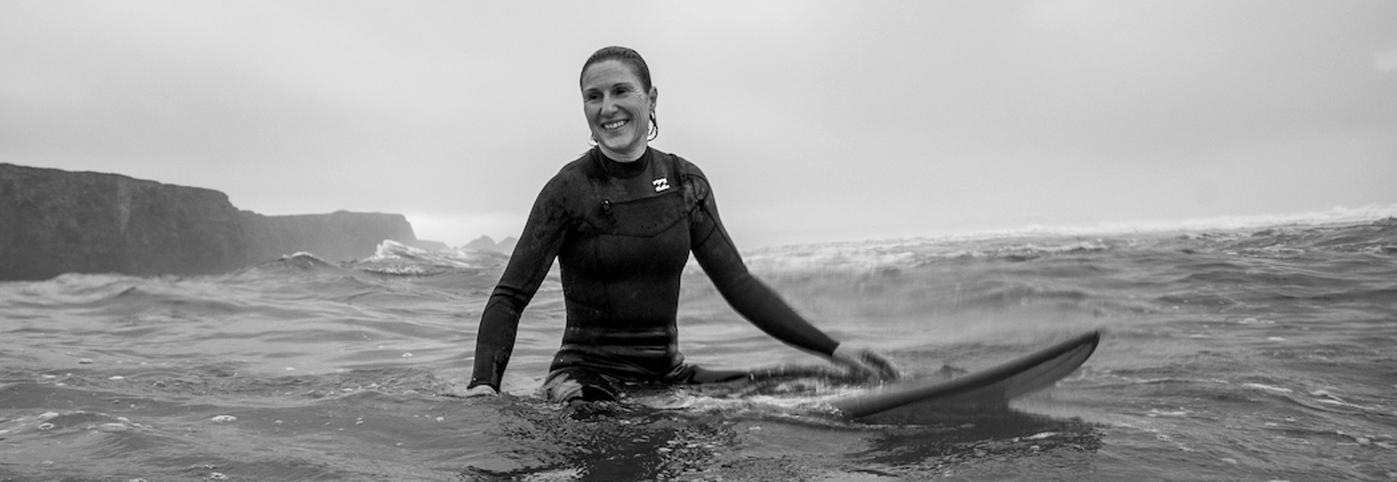 A black and white photograph showing a female surfer sitting on their board in the water, smiling.