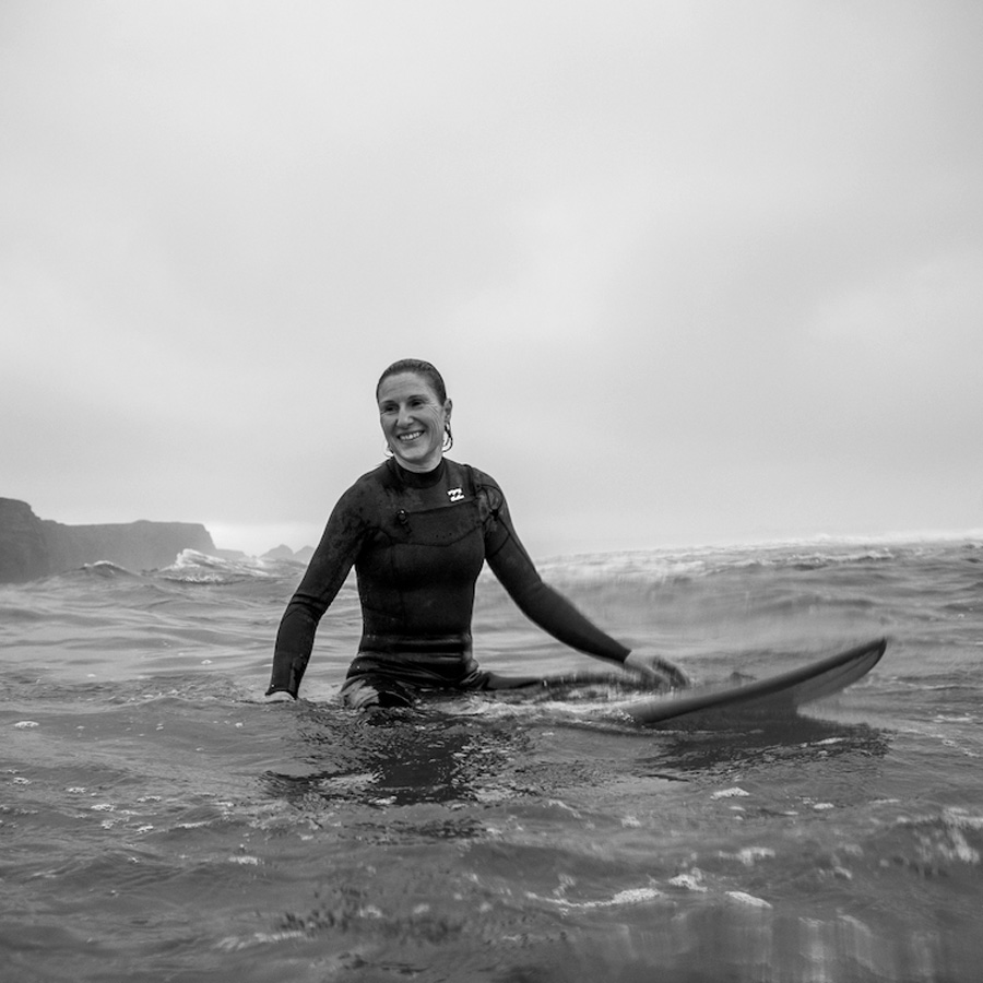 A black and white photograph showing a female surfer sitting on their board in the water, smiling.