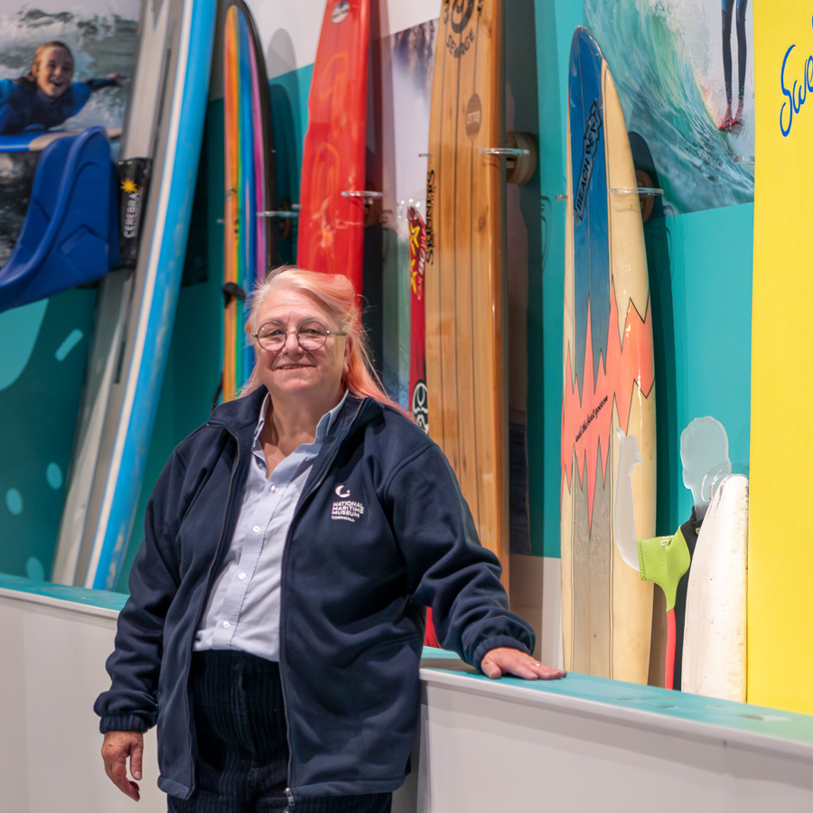 A colour photograph of a lady wearing a navy blue museum branded fleece, standing in front of a display of surfboards.