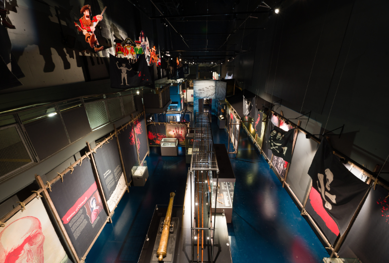 A photograph of an exhibition space, taken from above, with black and red banners, glass cases, a large metal cannon and graphics of pirate characters on display.