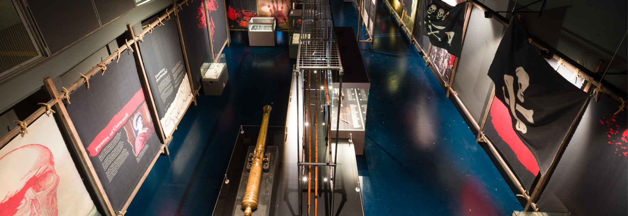 A photograph of an exhibition space, taken from above, with black and red banners and glass cases on display.
