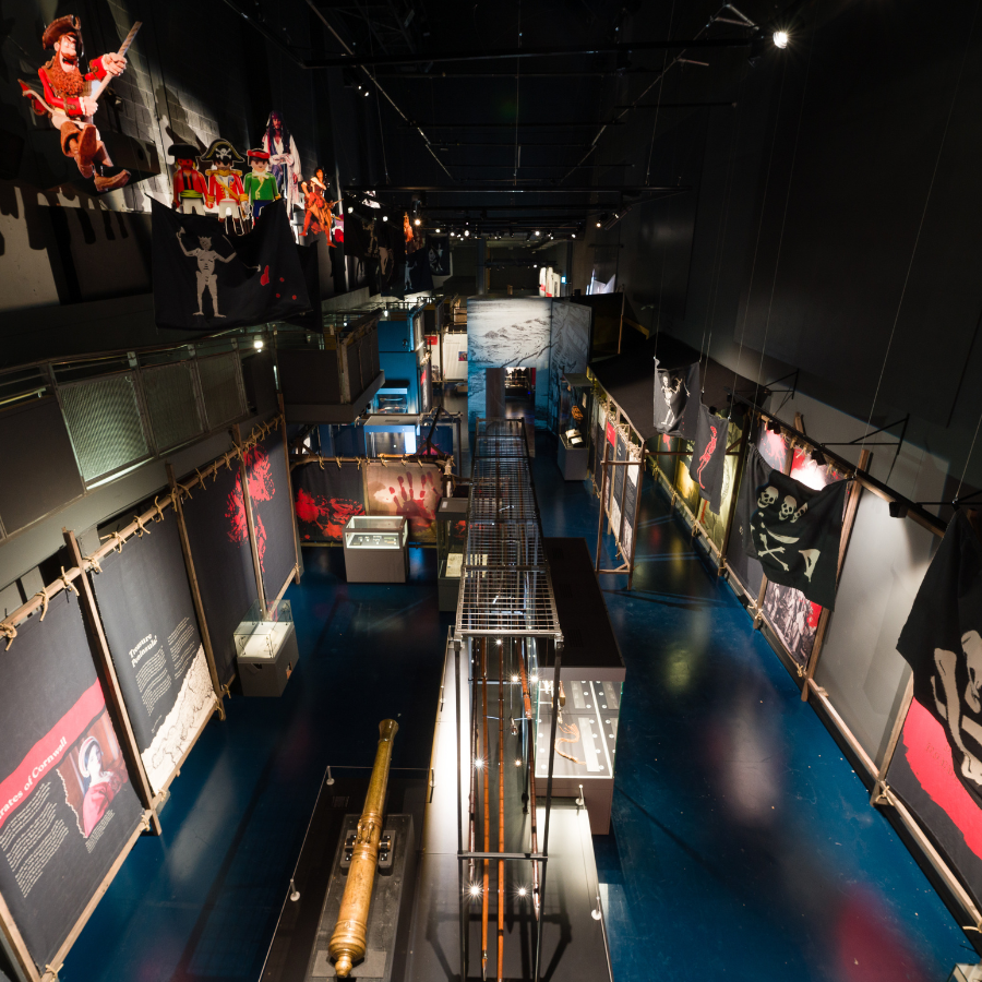 A photograph of an exhibition space, taken from above, with black and red banners, glass cases, a large metal cannon and graphics of pirate characters on display.