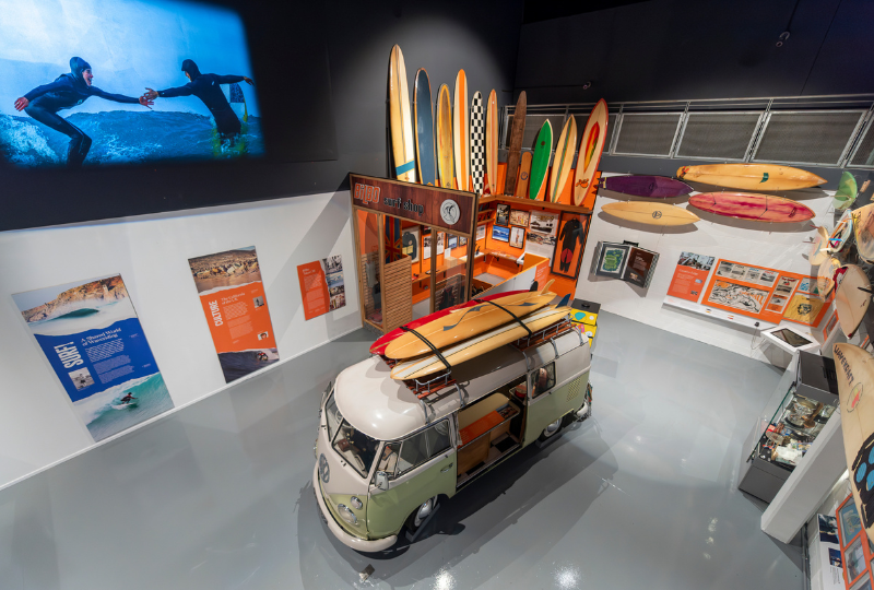 A photograph of an exhibition space, taken from above, a vintage VW campervan with surfboards attached to the roof is surrounded by surfboards lining the walls of the exhibition and colourful display panels. On one wall a projected surf film is playing.