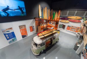 A photograph of an exhibition space, taken from above, a vintage VW campervan with surfboards attached to the roof is surrounded by surfboards lining the walls of the exhibition and colourful display panels. On one wall a projected surf film is playing.