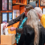 A man wearing a bright shirt and dark suit points to a photograph while talking to two people, he is surrounded by an exhibition of picture frames