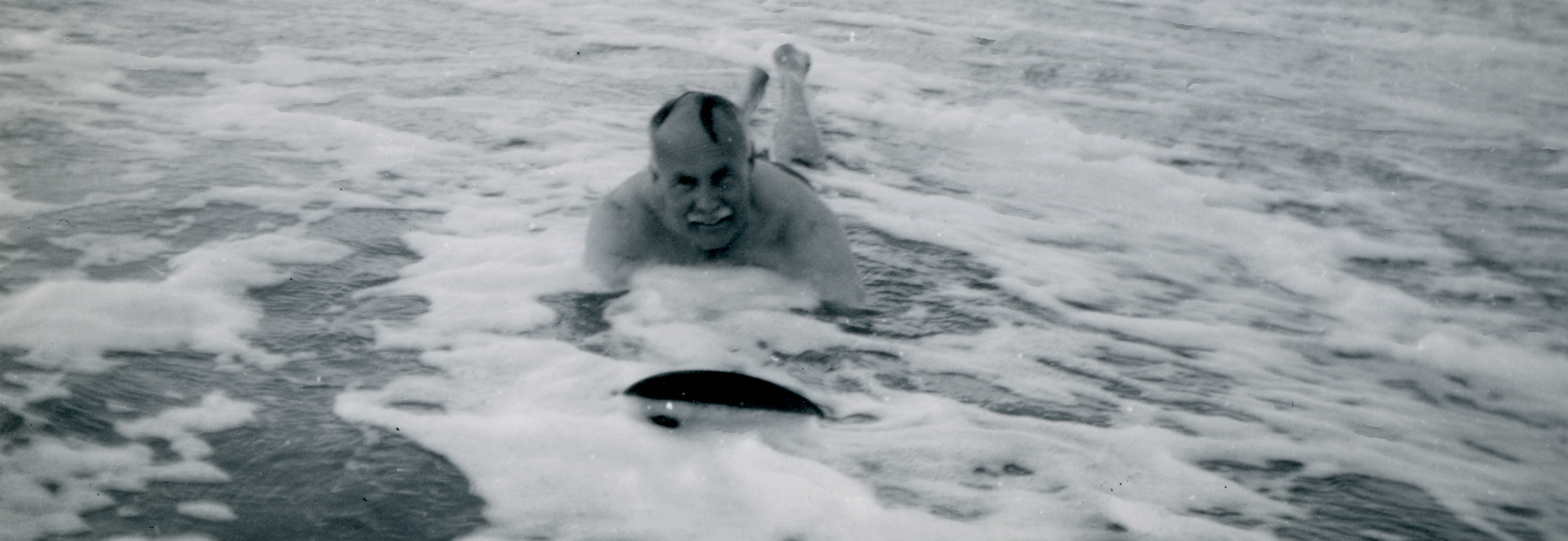 A black and white photograph of a man smiling as he is riding a wooden board into shallow water towards the camera, figures can be seen in the distance standing in shallow water.