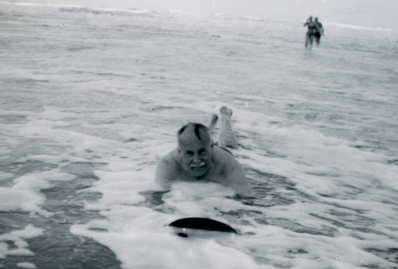 A black and white photograph of a man smiling as he is riding a wooden board into shallow water towards the camera, figures can be seen in the distance standing in shallow water.
