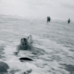 A black and white photograph of a man smiling as he is riding a wooden board into shallow water towards the camera, figures can be seen in the distance standing in shallow water.