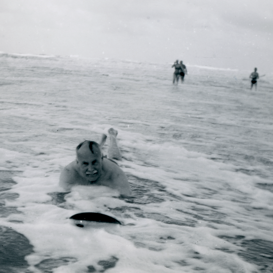 A black and white photograph of a man smiling as he is riding a wooden board into shallow water towards the camera, figures can be seen in the distance standing in shallow water.