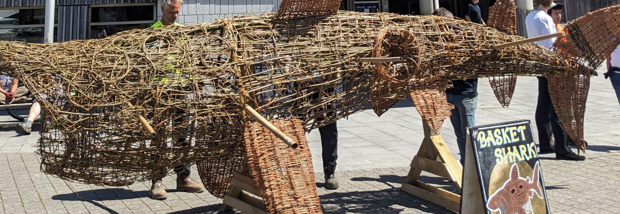 A large withy (willow) sculpture of a Basking Shark stands on a wooden base outside the National Maritime Museum Cornwall while people look on.