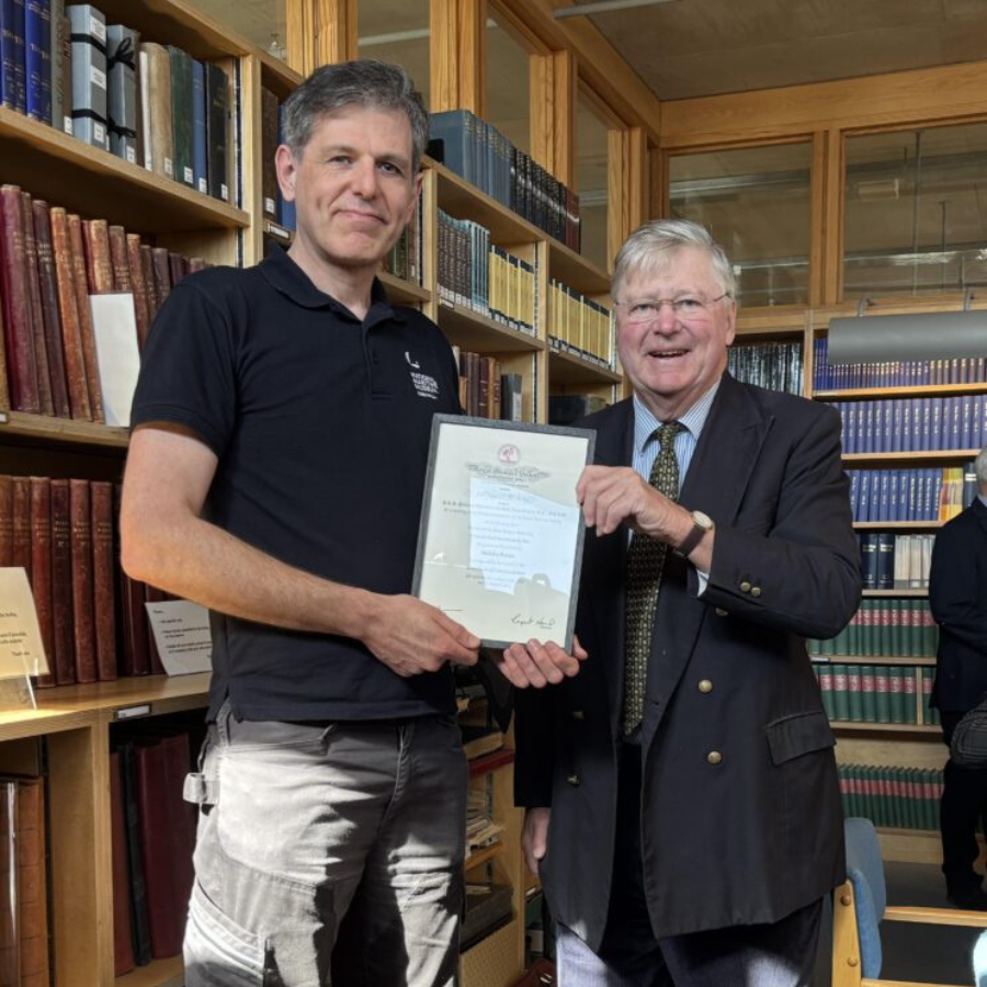 Two men are standing in a library, smiling at the camera, while both holding a framed certificate.