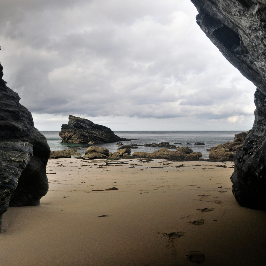 A photograph taken from inside a beach cave, looking out to the sea and large rocks in the distance and a trail of footprints in the sand in the foreground.