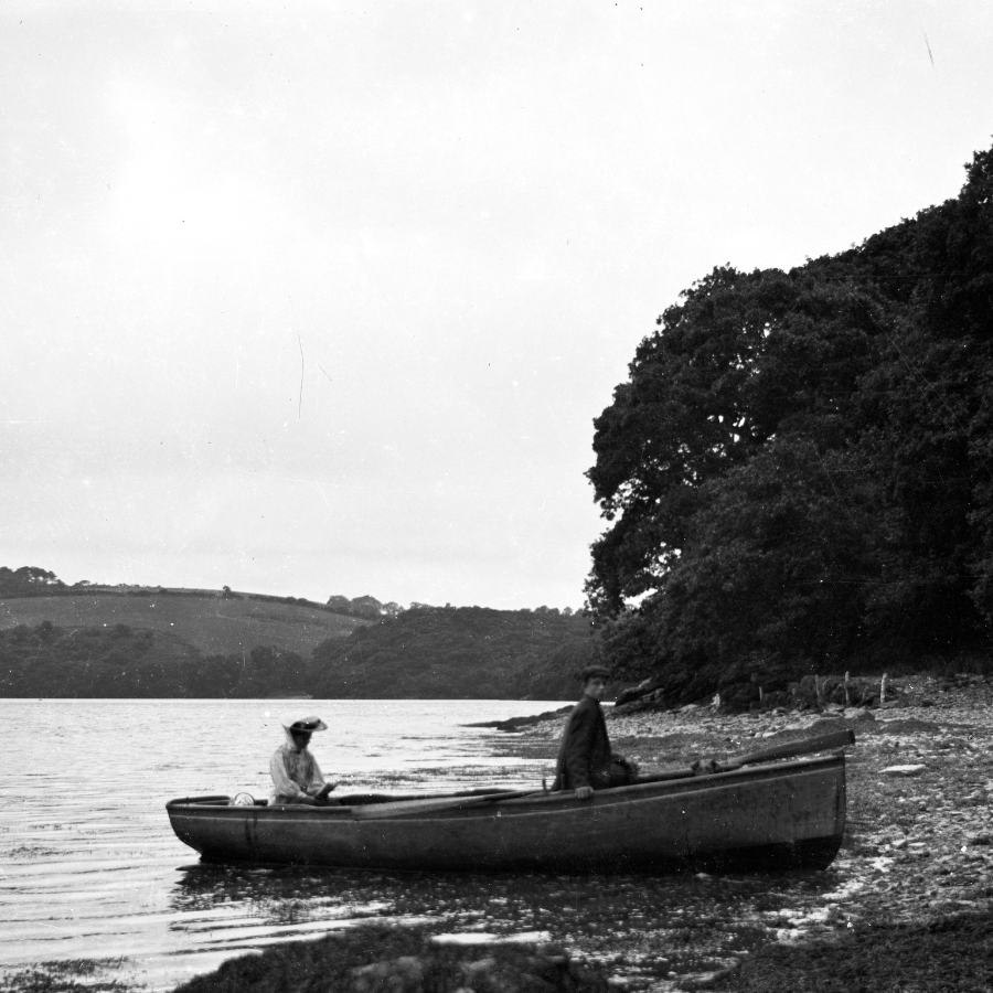 A black and white archive image shows two people in a small rowing boat arriving at the shore of a stony beach.