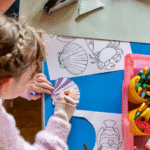 A photograph shows a young girl colouring-in a shell illustration, surrounded by craft materials.