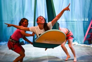 In this photo there are three performers. One performers is laying on a yellow surfboard which is suspended in the air. The two other performers are either side of the person on the yellow surfboard.
