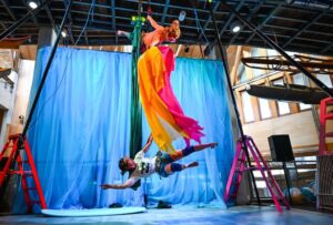 Two aerial dancers are suspended from a metal frame, with a blue cloth background behind them. 