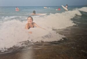 Archive photograph of a young girl riding a bodyboard in shallow waves, she is smiling at the camera.