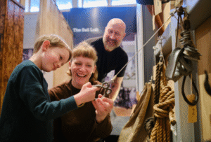 A family enjoying a day out at National Maritime Museum Cornwall, practicing knot-tying with ropes and nautical objects hanging from a wall.