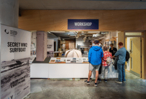 A photograph of Museum visitors looking into the boatbuilding workshop while the Museum's Boat Collection Manager explains the latest build project, which can be seen behind him.