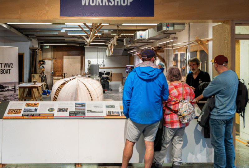 A photograph of Museum visitors looking into the boatbuilding workshop while the Museum's Boat Collection Manager explains the latest build project, which can be seen behind him.