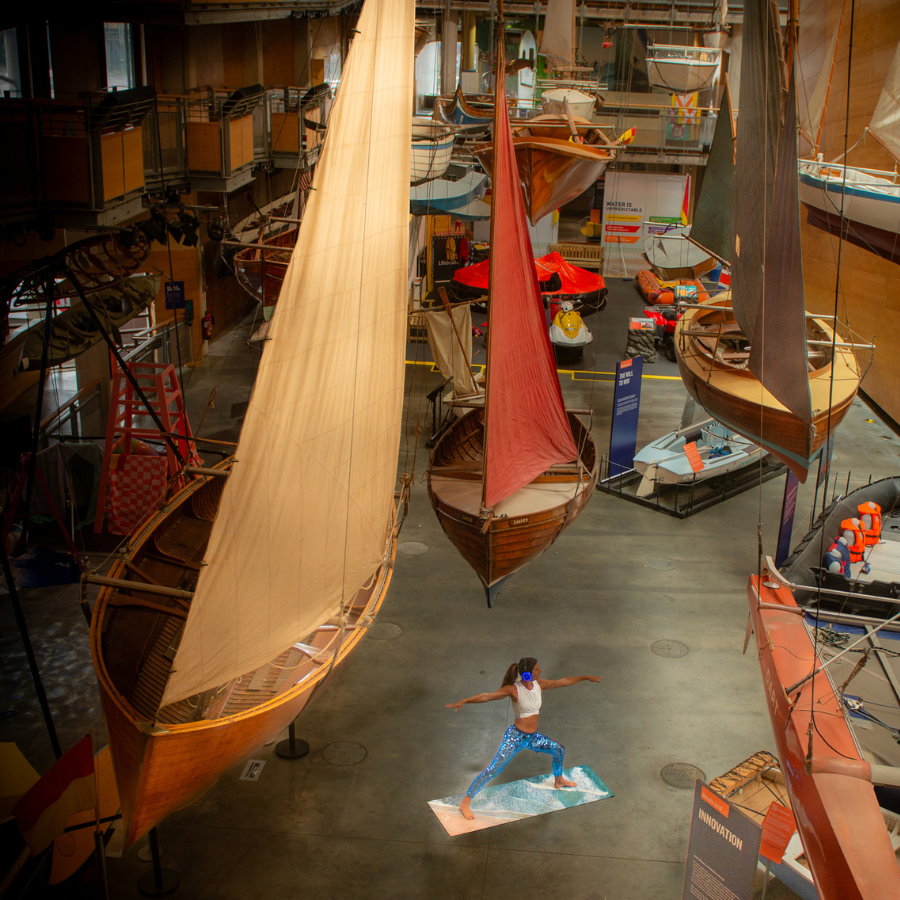 A woman stands in a yoga post surrounded by boats in the Boat hall at National Maritime Museum Cornwall