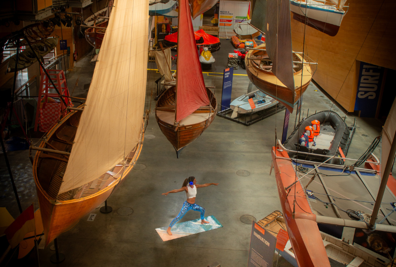 A woman stands in a yoga post surrounded by boats in the Boat hall at National Maritime Museum Cornwall