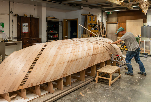A member of the boatbuilding team working on the Helford SOE Replica Surfboat build while the boat is upturned in the workshop.