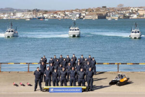 A group of Royal Navy personnel stand on the shore with three boats in the background.