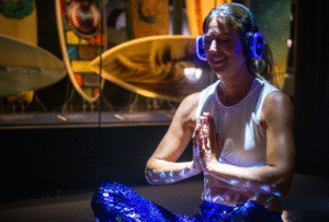 A woman sits in a yoga pose, wearing lit-up headphones surrounded by surfboards in the SURF! Exhibition at National Maritime Museum Cornwall