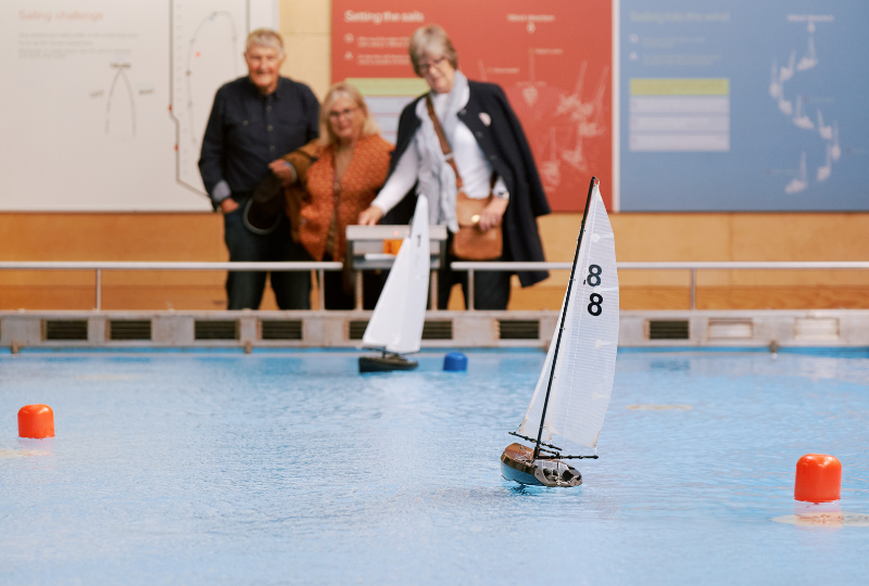 Two women and a man stand by a controller, maneuvering a miniature yacht on a boat pool.