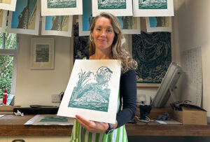 A photo of a women holding a linocut print with more prints hanging from a drying line behind her.