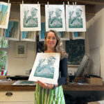 A photo of a women holding a linocut print with more prints hanging from a drying line behind her.