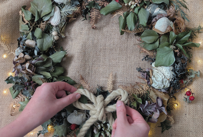 A close-up of hands adjusting a rope bow on a Coastal Christmas wreath of natural foliage.