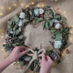 A close-up of hands adjusting a rope bow on a Coastal Christmas wreath of natural foliage.